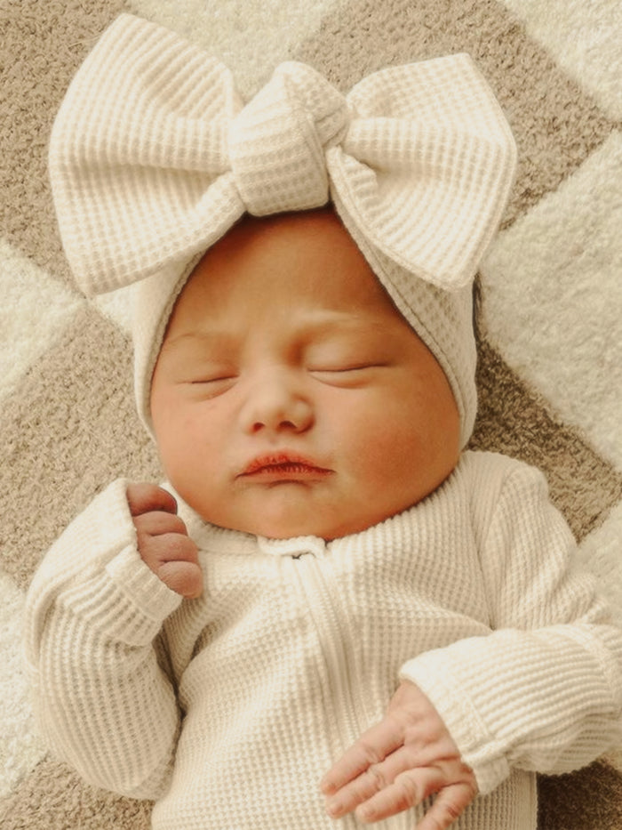 Newborn baby girl asleep in a cozy white outfit with a large bow headband on a textured blanket.