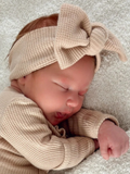 Newborn baby sleeping on a soft blanket, wearing a beige outfit and headband with a large bow.