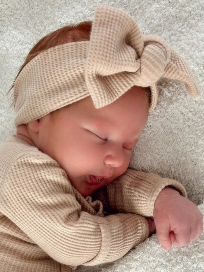 Newborn baby sleeping on a soft blanket, wearing a beige outfit and headband with a large bow.