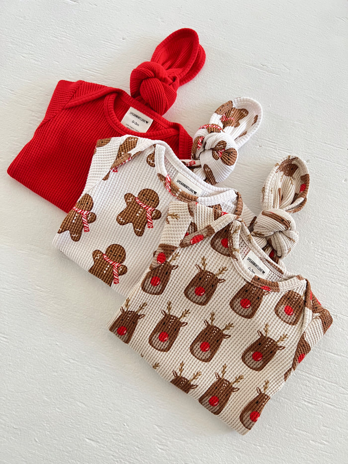 Three festive baby outfits: red long-sleeve top, white and tan reindeer, and gingerbread print knotted tops.