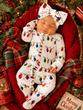 Baby in festive outfit with Christmas lights, sleeping peacefully on a red blanket surrounded by greenery and holiday decor.