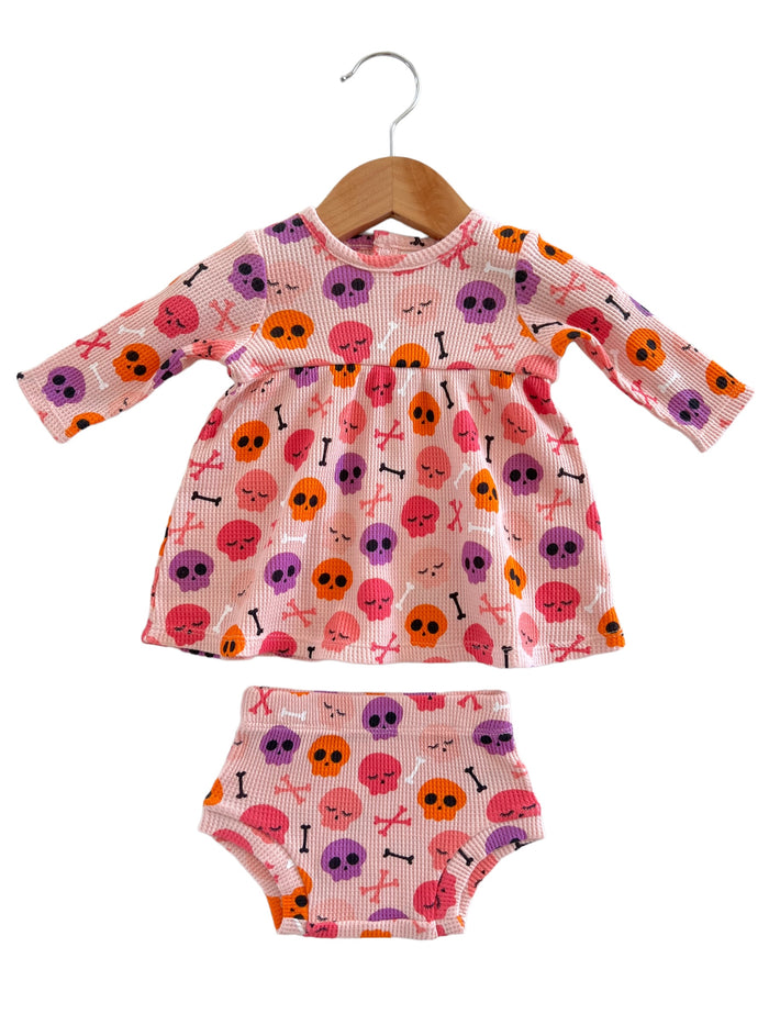 Pink dress and bloomers featuring colorful skull patterns, hanging on a wooden hanger. Perfect for kids' costumes.