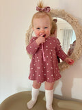 Smiling toddler in a heart-patterned dress holding a pink toothbrush, standing by a decorative mirror.