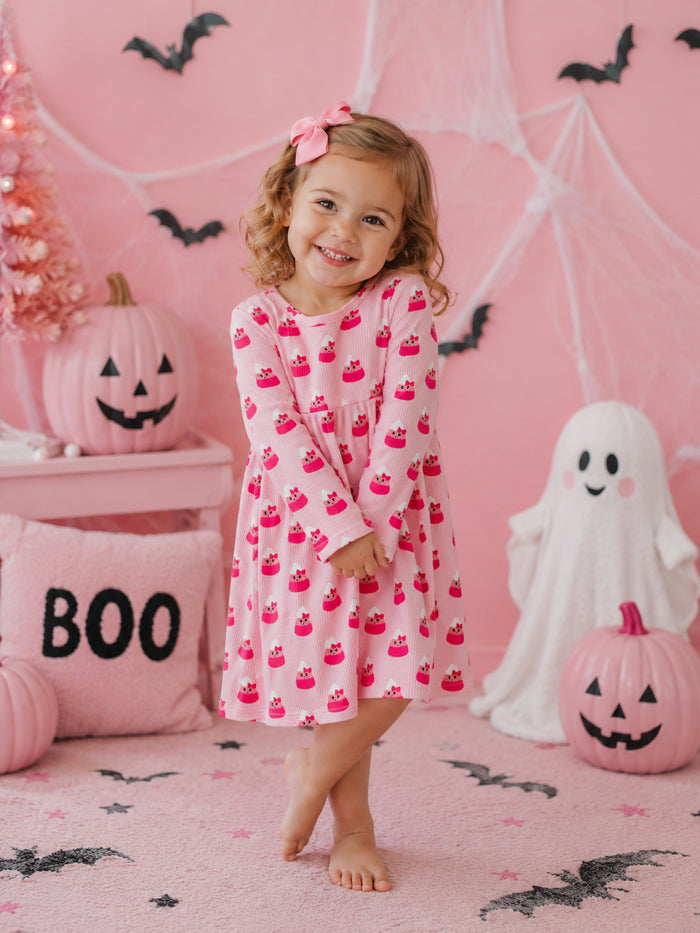 Smiling child in pink cupcake dress stands in a festive Halloween setting with pumpkins and decorations.