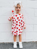 Smiling child in a heart-patterned dress, holding a heart-shaped lollipop against a white wall.