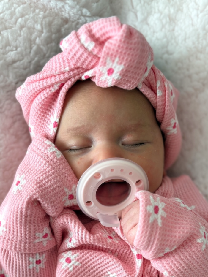 Baby sleeping peacefully with a pink headband and pacifier, wrapped in a cozy pink blanket with floral patterns.