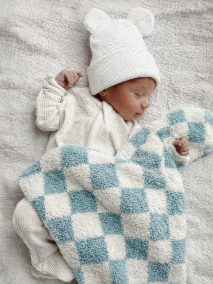 Newborn baby wearing a white bear hat, peacefully sleeping under a blue and white checkered blanket on a textured surface.