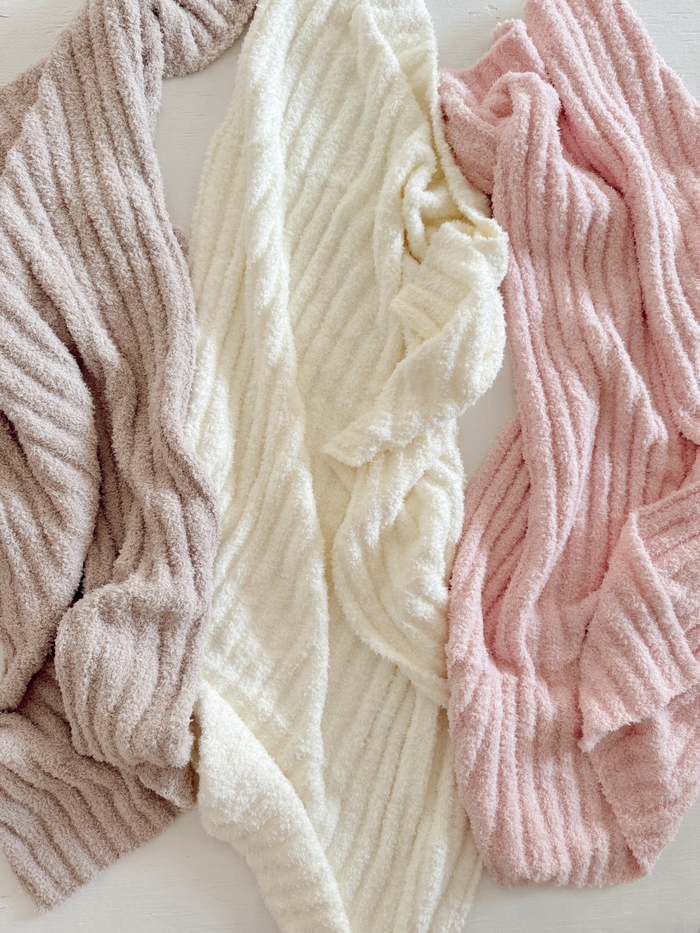 Three cozy, textured blankets in beige, white, and pink, arranged on a light surface.