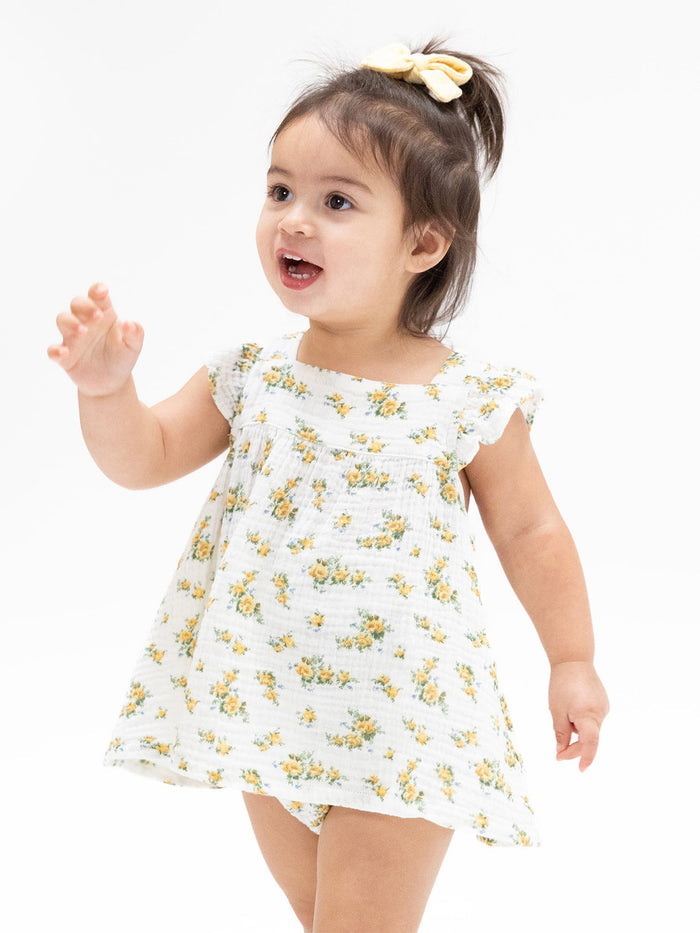 Toddler girl with brown hair in a yellow bow, wearing a floral dress, smiling while playing on a white background.