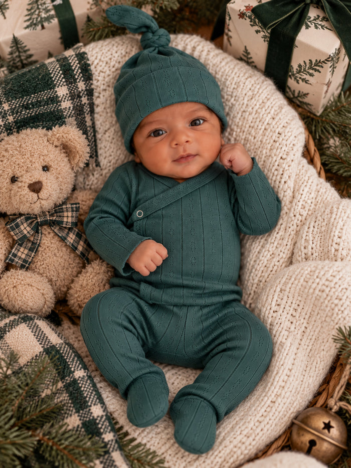 Infant in a green outfit with a matching hat, cuddling a teddy bear among holiday decor and wrapped gifts.