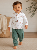 Smiling toddler in a white patterned shirt and green cargo pants, walking on a woven rug in a cozy setting.