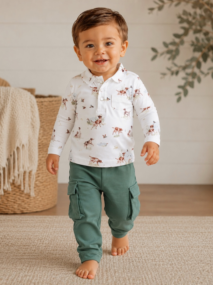 Smiling toddler in a white patterned shirt and green cargo pants, walking on a woven rug in a cozy setting.