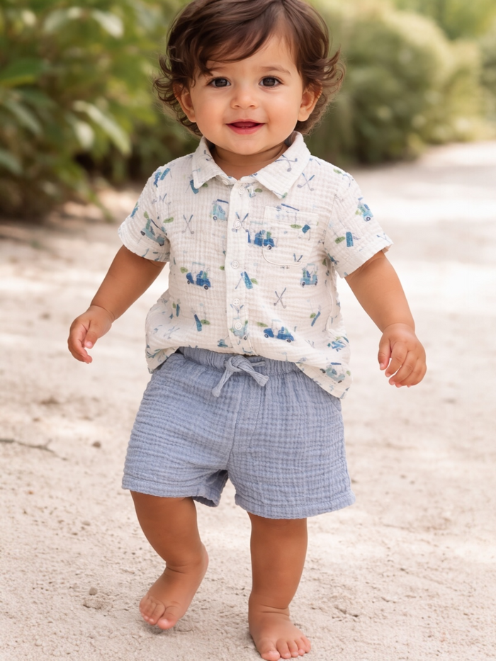 Smiling toddler in a patterned shirt and blue shorts walking on a sandy path surrounded by greenery.
