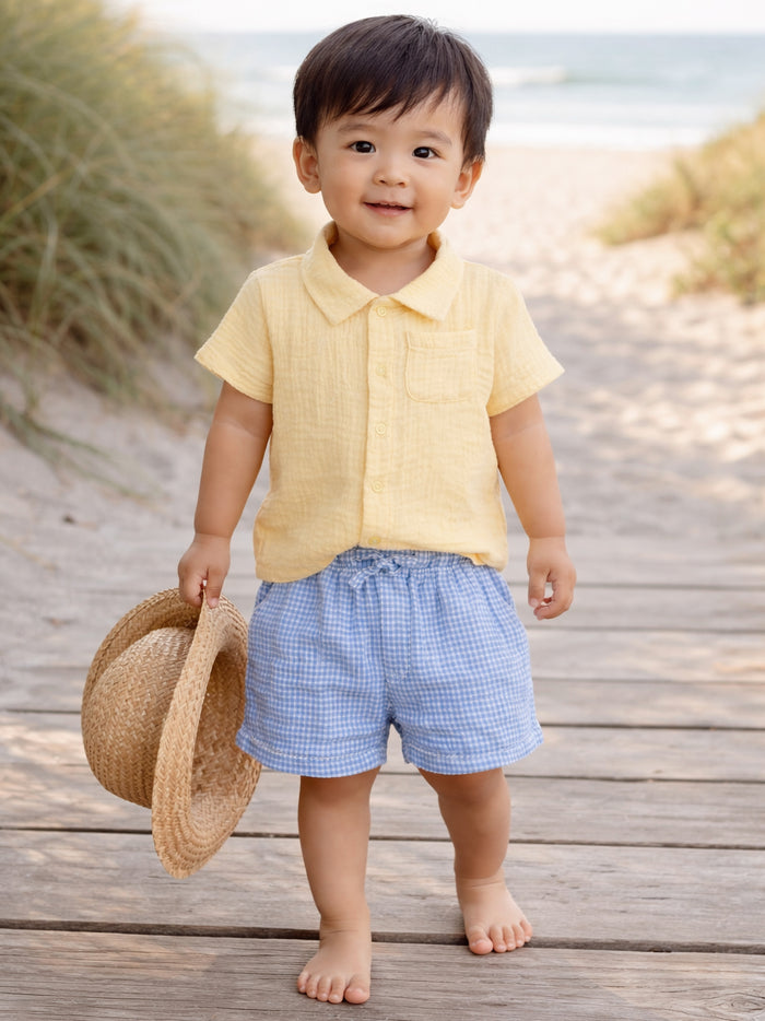 Smiling toddler in yellow shirt and blue shorts, holding a straw hat on a beach path.
