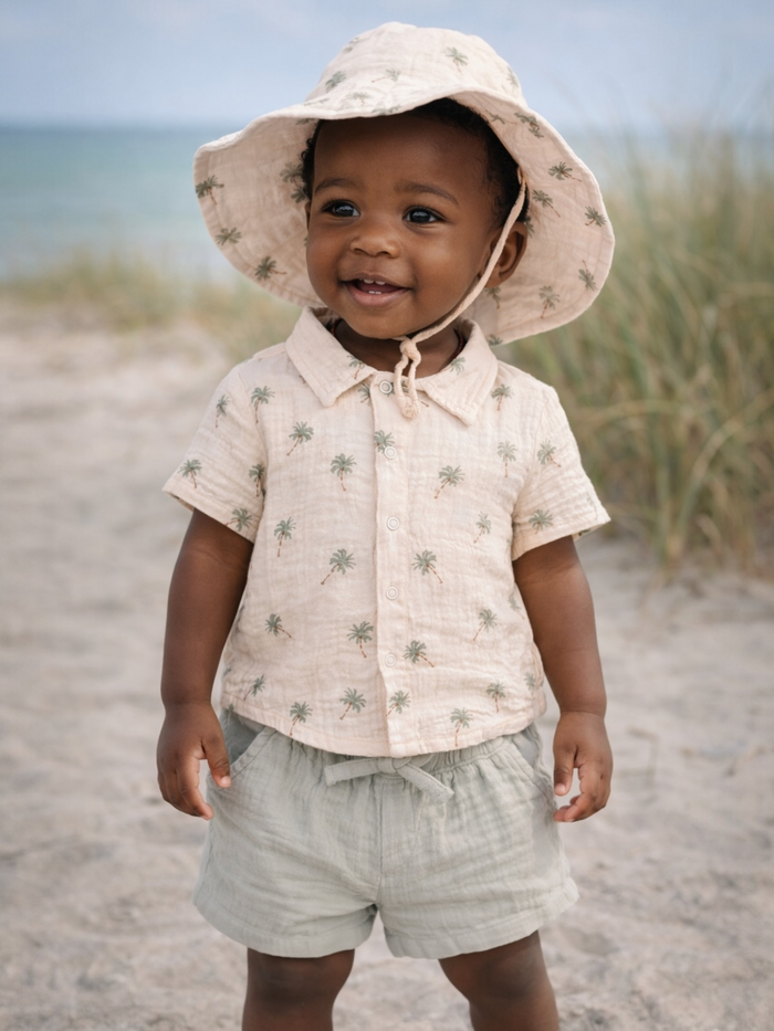 Smiling child in a palm-patterned shirt and hat at the beach, with grass and ocean in the background.