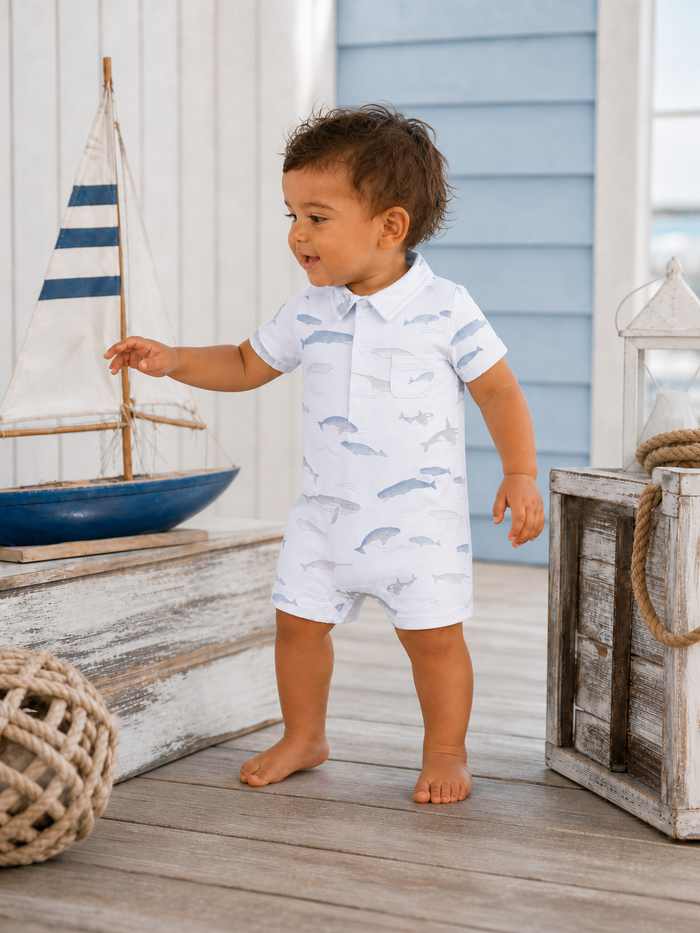 Toddler in a fish-patterned outfit stands near a sailboat on a wooden deck, smiling joyfully.