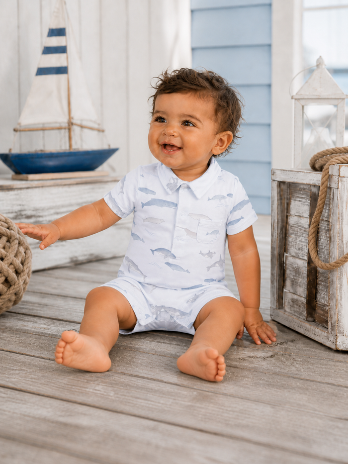 Smiling toddler in a fish-patterned outfit sitting on wooden flooring near nautical decor.
