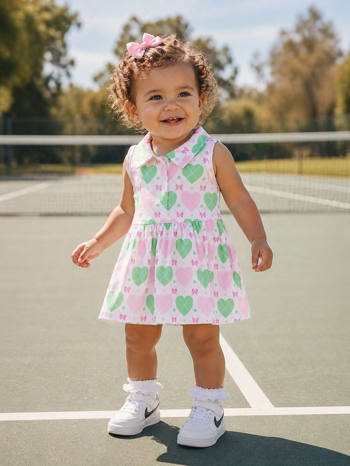 Smiling toddler in a heart-patterned dress and shoes, standing on a tennis court with trees in the background.