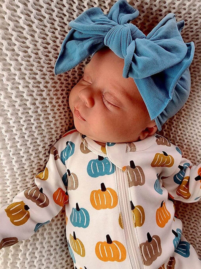 Sleeping baby in a pumpkin-patterned onesie, wearing a large blue bow headband, resting on a textured blanket.