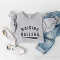 Gray sweatshirt with "Raising Ballers" text, paired with jeans and white sneakers on a neutral background.