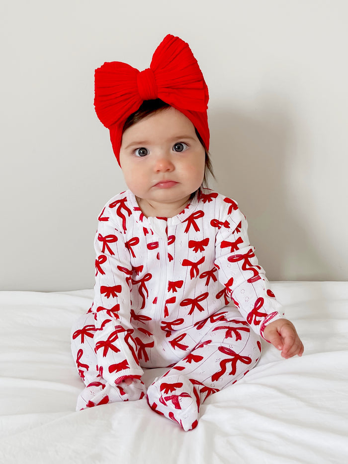 Infant girl in a white outfit with red bows, wearing a large red bow headband, seated on a white bed.