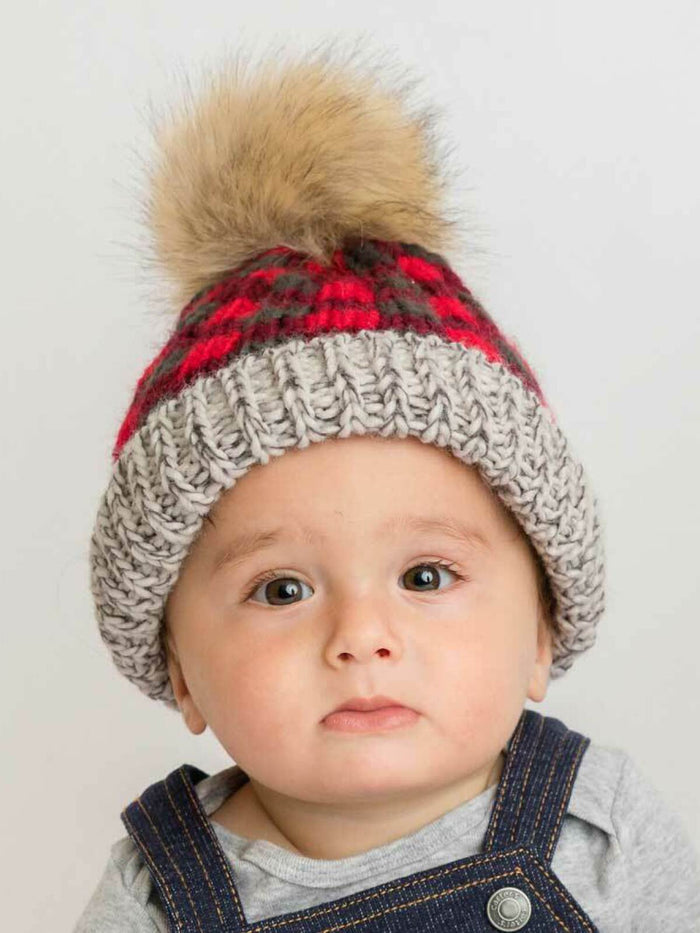 Baby in a knitted hat with a faux fur pom-pom, wearing a gray top and denim overalls, looking at the camera.