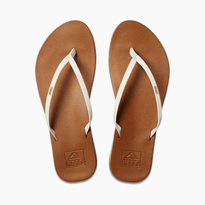 A pair of Reef Cushion Slim Flip Flops with white straps and gold embroidery on a brown vegan leather base.