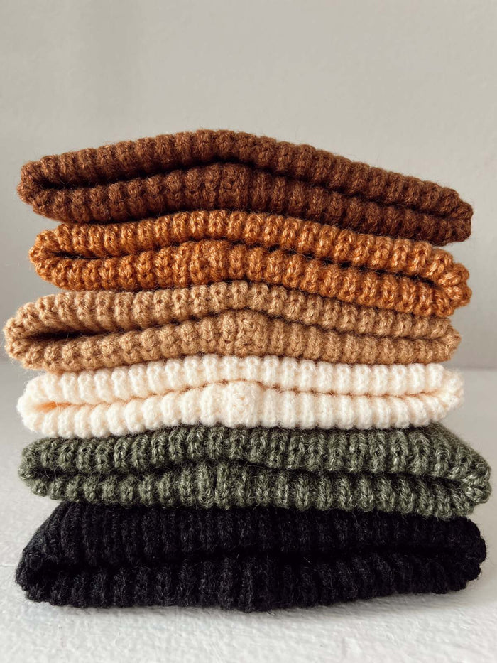 Stack of six folded knit scarves in shades of brown, cream, green, and black against a neutral background.