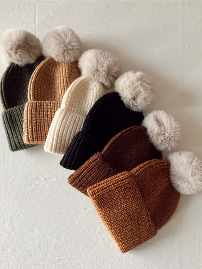Collection of knit beanies in various colors with pom-poms, arranged on a light background.