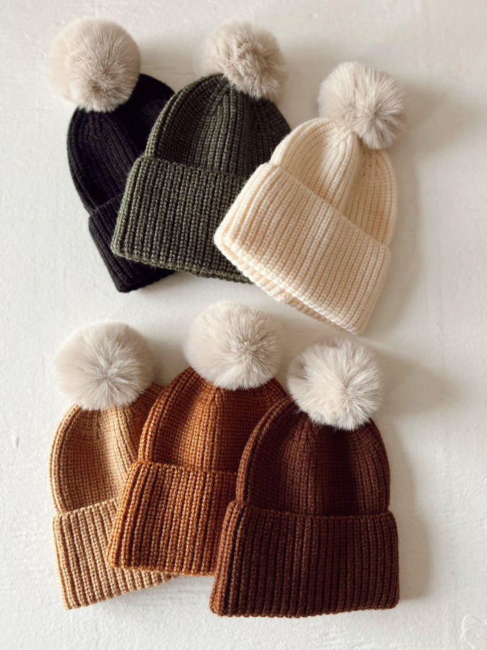 A collection of knitted beanies in black, olive, cream, tan, and brown with round faux fur pom poms on top.