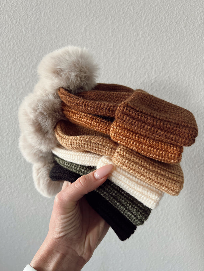 Hand holding stack of colorful knitted hats with faux fur pom-poms against a light background.