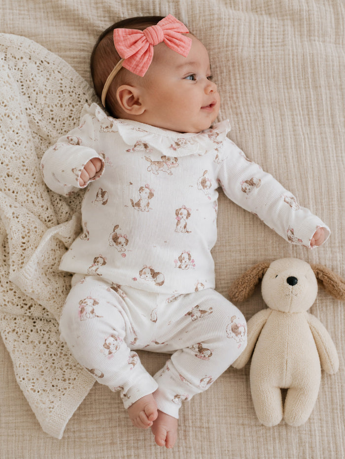 Infant in a patterned outfit with a pink bow, lying next to a soft toy on a textured blanket.