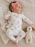 Infant in a patterned outfit with a pink bow, lying next to a soft toy on a textured blanket.