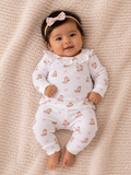 Smiling baby in a cozy outfit with a bear pattern and a pink bow headband, posed on a textured blanket.