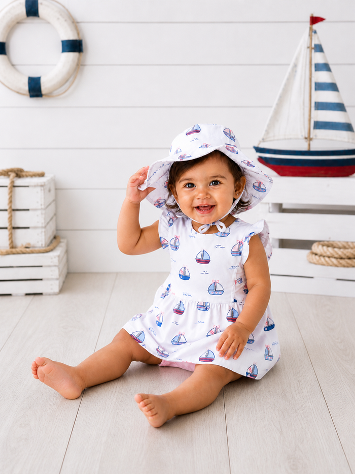 Smiling toddler in a boat-patterned dress and hat, seated on a wooden floor with nautical decor in the background.