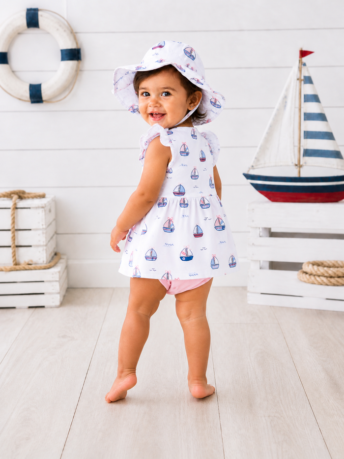 Smiling toddler in a sailboat-patterned dress and hat, standing near nautical decor in a bright, airy setting.