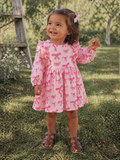 Smiling toddler in a pink dress with bows stands on grass, wearing brown sandals, surrounded by greenery.