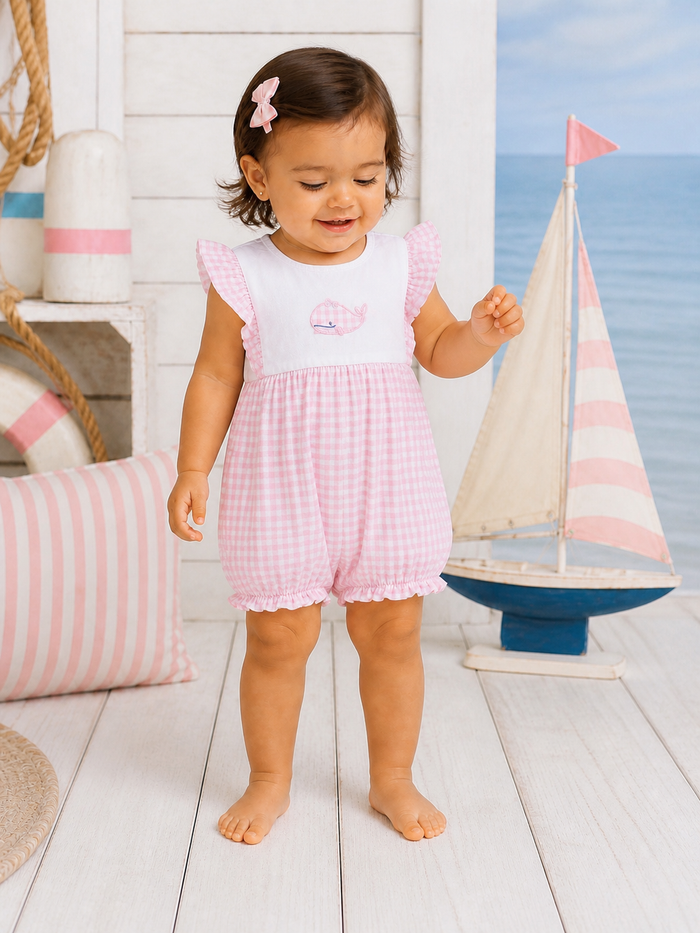 Toddler in a pink gingham outfit smiles near a toy sailboat and striped pillows by a beach-style backdrop.