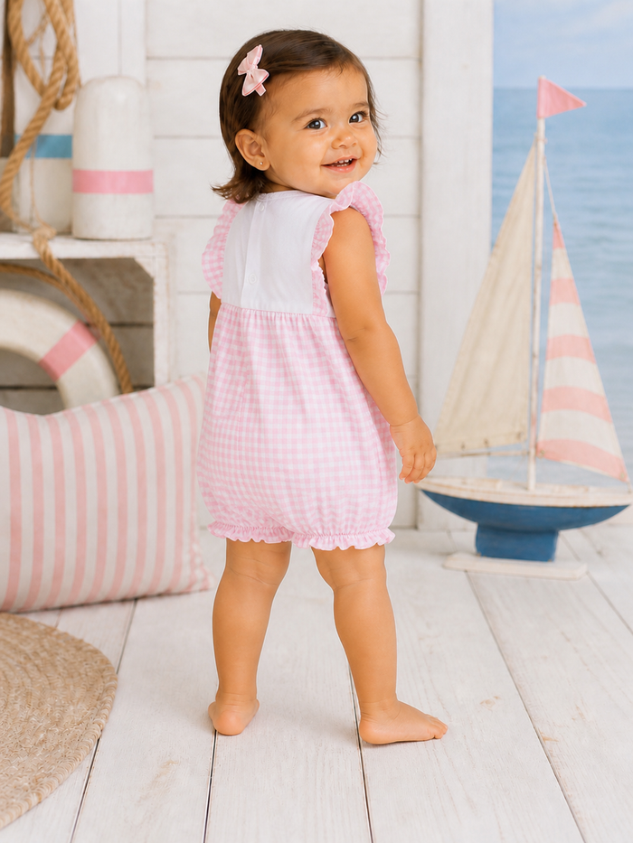 Smiling toddler in a pink gingham dress stands barefoot, facing away, near nautical decor and a toy sailboat.