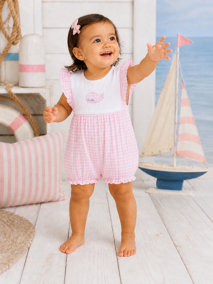 Smiling toddler in pink gingham romper reaching out, with nautical decor and a small sailboat in the background.