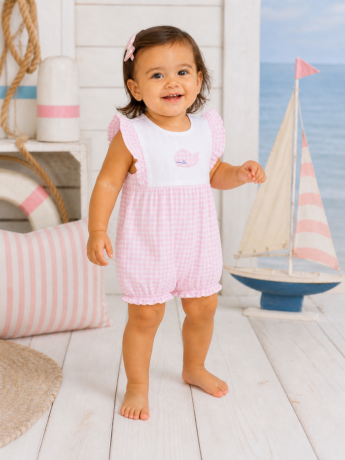 Smiling toddler in pink gingham outfit stands on wooden floor with nautical decor and toy sailboat background.