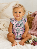 Smiling baby in floral onesie sitting on cozy blanket, surrounded by flowers and a "6 months" sign.