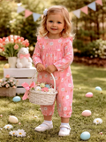 Smiling toddler in a pink bunny-patterned outfit holding a basket of pastel eggs in a spring garden setting.