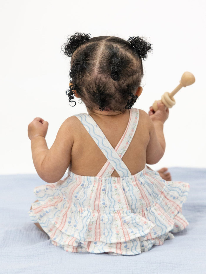 Baby girl with curly hair wearing a patterned dress, sitting and holding a wooden toy, seen from the back.