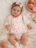 Smiling baby girl in a peach-patterned outfit, wearing a bow, surrounded by peaches and flowers. Soft, cozy setting.