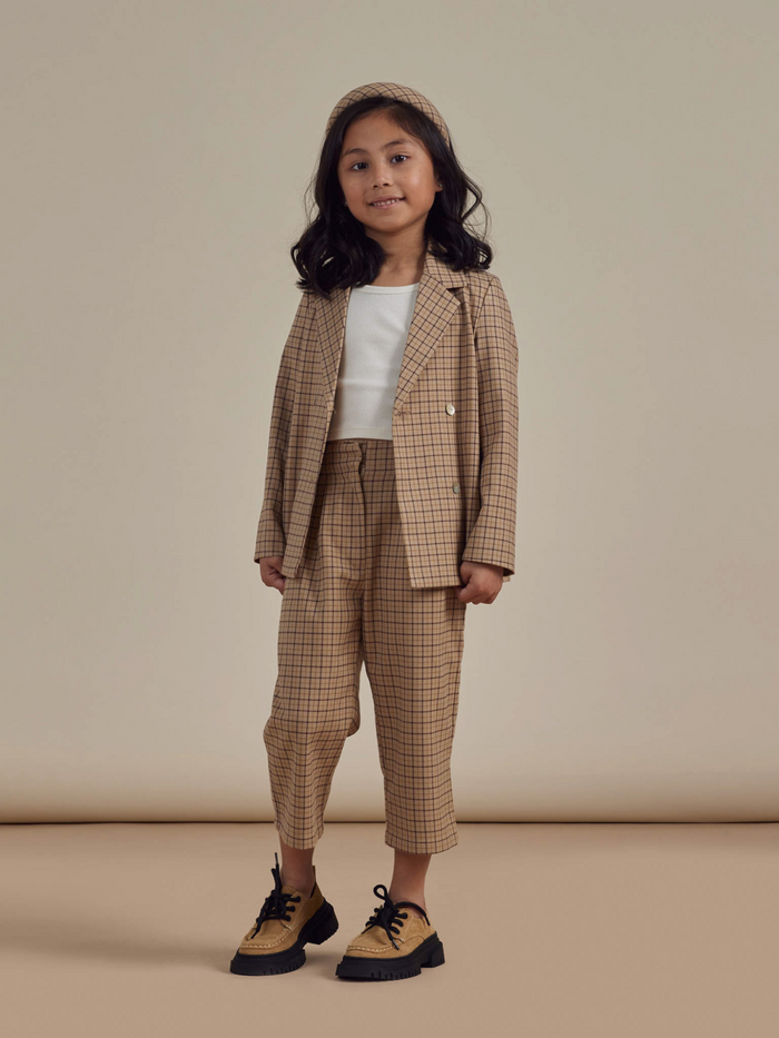Smiling child in a beige plaid suit with a white top and black shoes, standing against a neutral background.