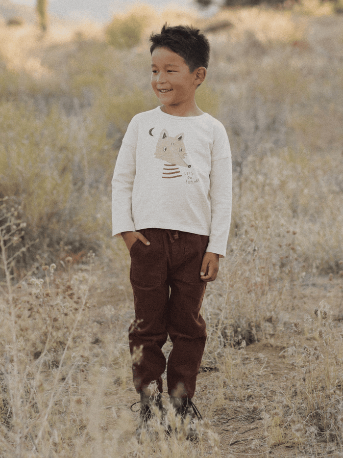 Smiling boy in a nature setting, wearing a t-shirt with a fox design and brown pants, surrounded by tall grass.