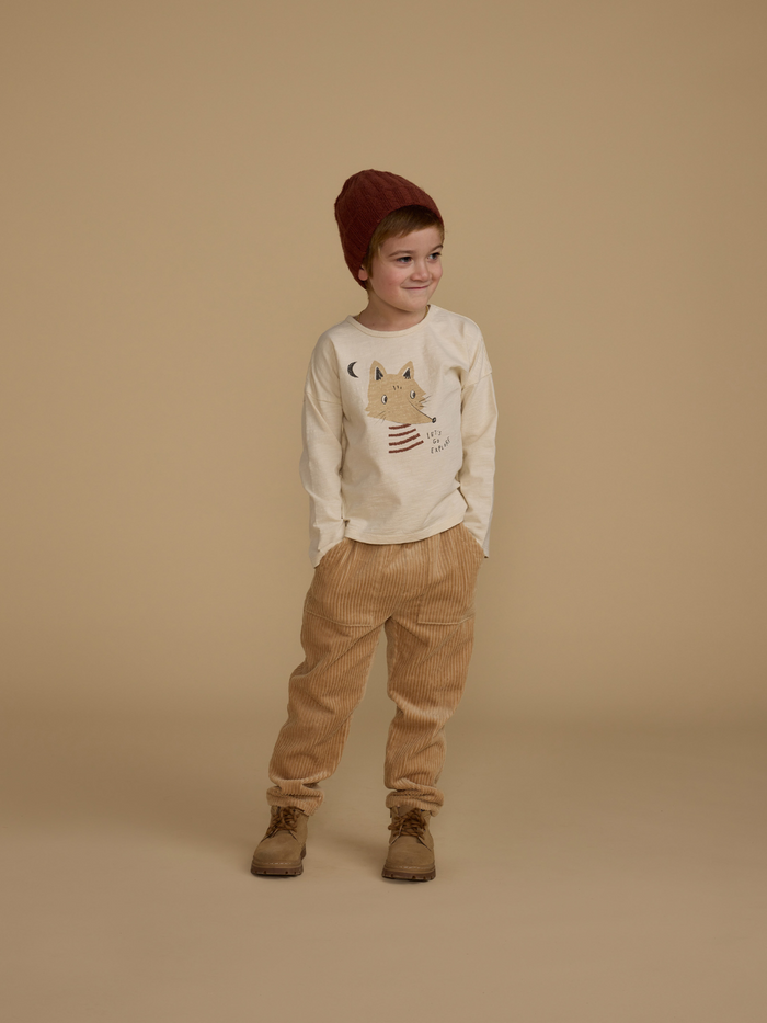 Child wearing a beige shirt with a fox design, tan corduroy pants, and a brown knit hat against a neutral background.