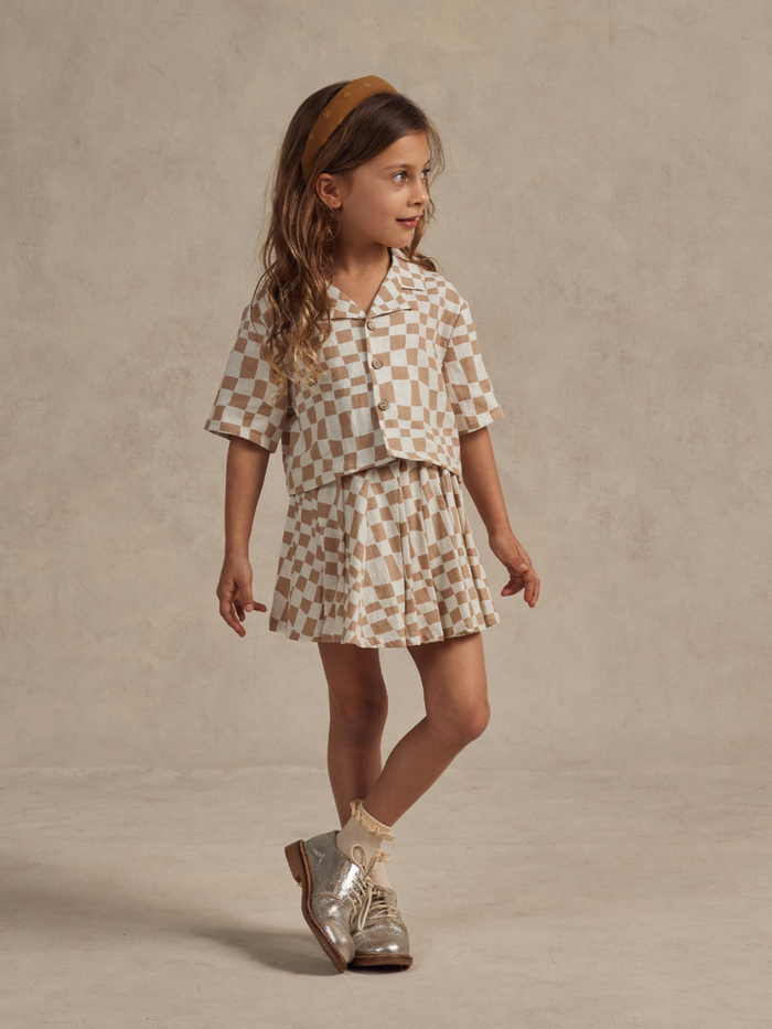 Child in a checked outfit with a matching headband, silver shoes, against a neutral background.