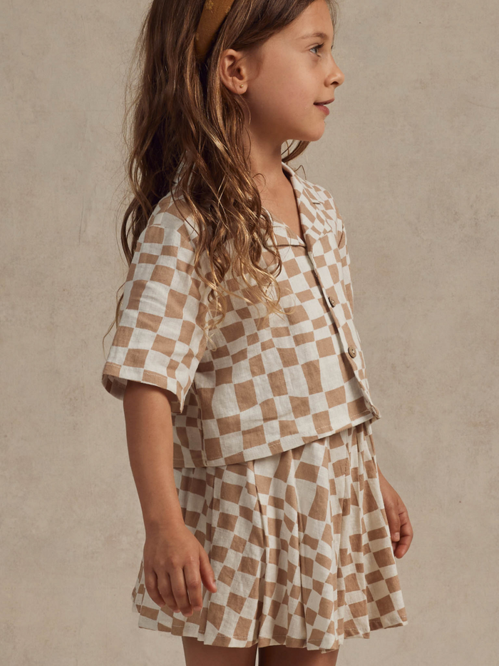 Girl in a checkered outfit with wavy hair, smiling softly against a neutral background.